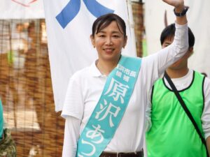 Atsumi Harasawa mit türkisfarbener Wahlkampfschärpe winkt lächelnd bei einer Straßenrede