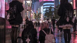 Jugendliche in schwarzer Rüschenkleidung mit Rucksäcken in einer nassen, bunt beleuchteten Straße in Kabukichō, von hinten