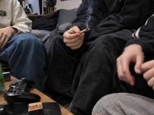 Junge Menschen sitzen auf einem Sofa, einer hält einen brennenden Joint, auf dem Tisch liegen ein Gamecontroller und Zigarettenpapier