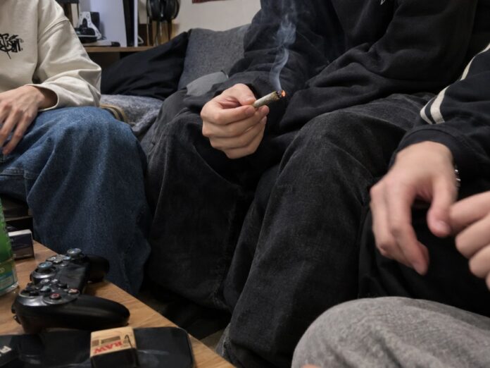 Junge Menschen sitzen auf einem Sofa, einer hält einen brennenden Joint, auf dem Tisch liegen ein Gamecontroller und Zigarettenpapier