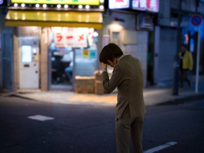 Ein Mann im Anzug steht nachts allein auf einer Straße in Tokio, die Hand an der Stirn