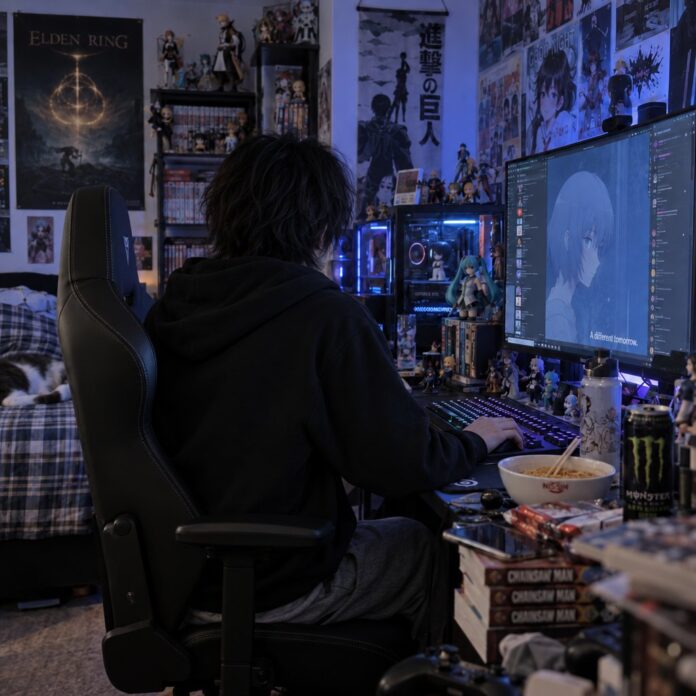 Junger Mann von hinten an seinem Computer in einem abgedunkelten Zimmer