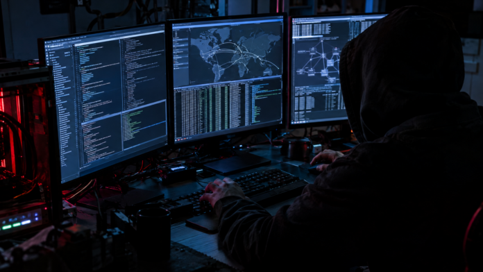Vermummter Hacker an mehreren Bildschirmen mit Code und Weltkarte