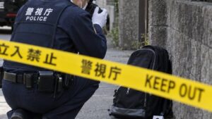Ein japanischer Polizist fotografiert hinter einem Absperrband einen Rucksack am Straßenrand, im Hintergrund ein Streifenwagen