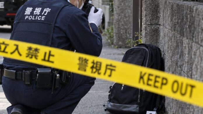 Ein japanischer Polizist fotografiert hinter einem Absperrband einen Rucksack am Straßenrand, im Hintergrund ein Streifenwagen