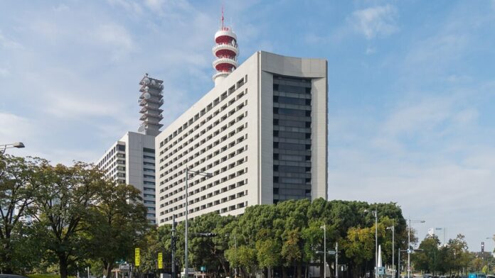 Hochhaus des Hauptquartiers der Tokioter Polizei mit rot-weißem Funkturm auf dem Dach, davor Bäume und eine Straßenkreuzung