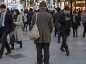 Ein älterer Mann mit Mütze und Stofftasche steht still in einer belebten Einkaufsstraße in Tokio, Passanten gehen an ihm vorbei