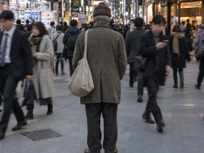 Ein älterer Mann mit Mütze und Stofftasche steht still in einer belebten Einkaufsstraße in Tokio, Passanten gehen an ihm vorbei