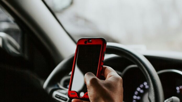 Symbolbild: Eine Hand hält ein Smartphone mit roter Hülle vor dem Lenkrad eines Autos