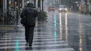 Eine Person in Regenkleidung überquert bei starkem Regen einen Zebrastreifen in einer japanischen Stadtstraße, im Hintergrund Autos mit Scheinwerferlicht