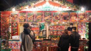 Besucher stehen abends vor einer beleuchteten Weihnachtsmarktbude mit deutschen Speisen