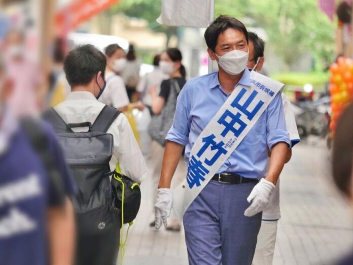 Takeharu Yamanaka mit Wahlkampfschärpe und Maske auf der Straße